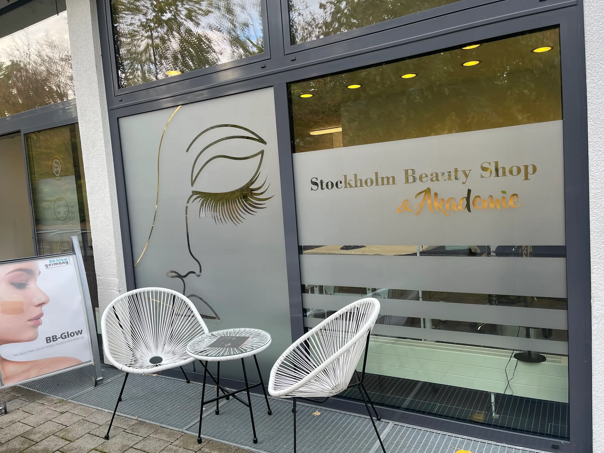 Schaufenster von Stockholm Beauty mit goldenem Gesicht als Fensterbild, Schriftzug und zwei weißen Stühlen davor